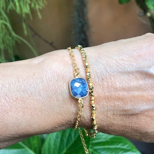 May include: Two gold bracelets are displayed. One features a square, faceted, blue gemstone set in a gold frame. The other is a delicate chain of small, gold, faceted beads. The bracelets are worn on a wrist.