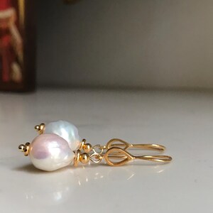 May include: A pair of gold earrings with teardrop-shaped hooks. Each earring features a large, lustrous, off-white baroque pearl with a hint of pink and gold accents. The earrings are displayed on a white surface.
