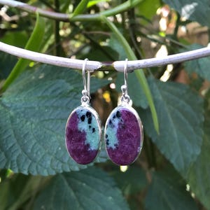 Op de afbeelding: Een paar ovale oorbellen met een zilveren zetting. Elke oorbel heeft een levendige kleurencombinatie: diep magenta, lichtblauwgroen en zwarte spikkels. De oorbellen hangen aan zilveren haken en tonen een natuursteenontwerp.