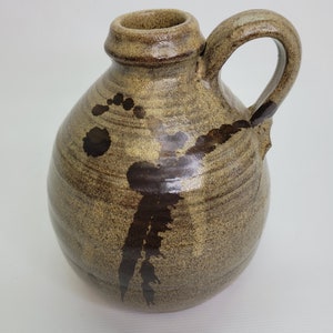May include: A handmade, brown ceramic jug with a rounded body and a handle. The jug features dark brown abstract designs and a textured surface. The opening is small, and the jug is suitable for decorative purposes.