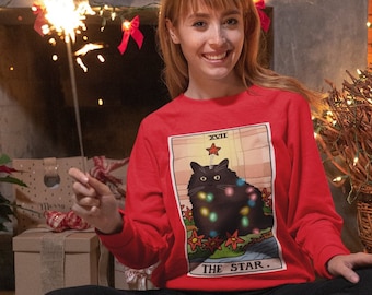 Może przedstawiać: Kobieta w czerwonym swetrze z długimi rękawami z ilustracją czarnego kota i napisem "XVII THE STAR." Kot jest otoczony gwiazdami i ma czerwoną gwiazdę na głowie. Kobieta siedzi na podłodze ze skrzyżowanymi nogami i trzyma iskrę.