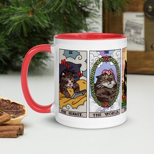 Puede incluir: Taza de cer&aacute;mica blanca con borde rojo. La taza presenta un dise&ntilde;o de cuatro cartas de tarot con gatos representados como los personajes. Las cartas son "El Ermita&ntilde;o", "El Mundo", "La Torre" y "La Estrella".