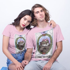 Pu&ograve; includere: Due persone indossano magliette rosa con un'illustrazione di un gatto in una ghirlanda, il testo "XXI THE WORLD" &egrave; sulla maglietta.