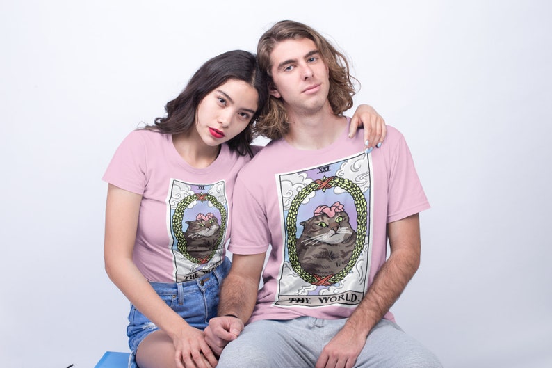 Pu&ograve; includere: Due persone indossano magliette rosa con un'illustrazione di un gatto in una ghirlanda, il testo "XXI THE WORLD" &egrave; sulla maglietta.