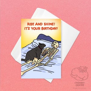 May include: A birthday card with a humorous illustration. The card features a skeleton in bed with a black cat, and the text "Rise and Shine! It's Your Birthday!" is displayed in orange and red.
