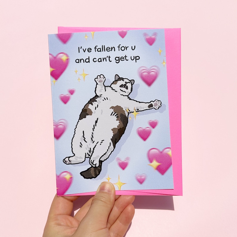 Puede incluir: Una tarjeta de felicitaci&oacute;n con un fondo azul claro que presenta un gato de dibujos animados con pelaje marr&oacute;n y blanco, rodeado de corazones rosas. La tarjeta dice "I've fallen for u and can't get up" y se sostiene frente a un sobre rosa.