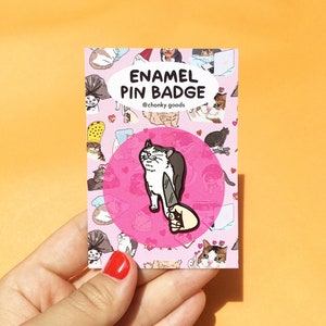 May include: A pink enamel pin badge featuring a white cat with black markings holding a knife. The cat is looking at the viewer with a surprised expression. The badge is surrounded by a pink circle with the text "Enamel Pin Badge" and "@chonky.goods" in black.