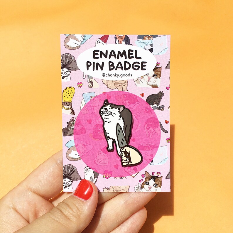 May include: A pink enamel pin badge featuring a white cat with black markings holding a knife. The cat is looking at the viewer with a surprised expression. The badge is surrounded by a pink circle with the text "Enamel Pin Badge" and "@chonky.goods" in black.