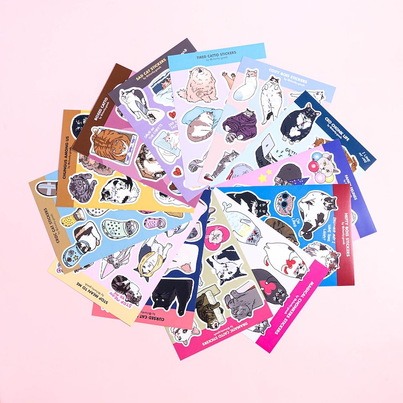 May include: A collection of 16 sticker sheets featuring various illustrations of cats in different poses and situations. The sticker sheets are colourful and have a variety of backgrounds, including pink, blue, yellow, and green. The sticker sheets are arranged in a fan shape on a pink background.