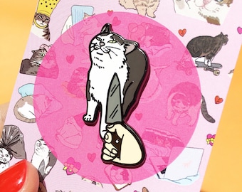 May include: A white and gray cat with a knife in its paw, holding the knife up in the air. The cat is standing on its hind legs. The image is a pin on a pink background with a circle around the cat. The background is covered with small images of cats.