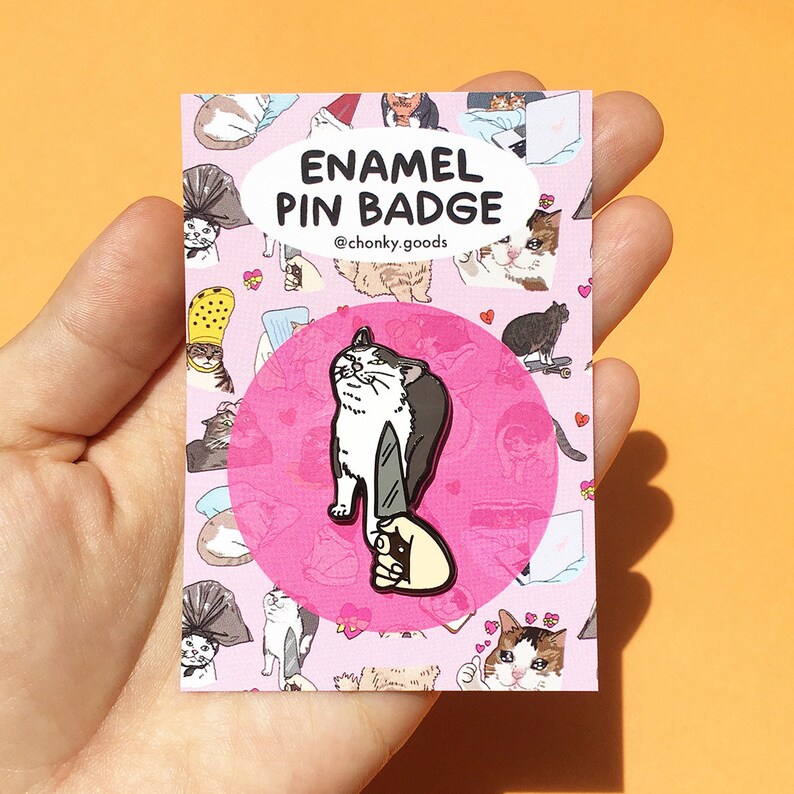 May include: A pink and white enamel pin badge featuring a white cat holding a knife. The text "ENAMEL PIN BADGE" and "@chonky_goods" are printed on the badge.