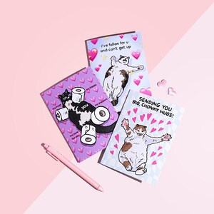 Puede incluir: Tres tarjetas de felicitaci&oacute;n ilustradas con gatos y texto. Una tarjeta dice "I've fallen for u and can't get up." Otra dice "Sending you big, chonky hugs!" Tambi&eacute;n se ve un bol&iacute;grafo rosa. La tercera tarjeta presenta un gato con rollos de papel higi&eacute;nico.