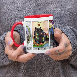 Puede incluir: Taza de cer&aacute;mica blanca con borde rojo, que presenta un gato negro con luces de colores, rodeado de un dise&ntilde;o floral. El gato est&aacute; representado en un estilo de carta de tarot con el texto "The Star" y el n&uacute;mero romano "XVIII" sobre el gato.