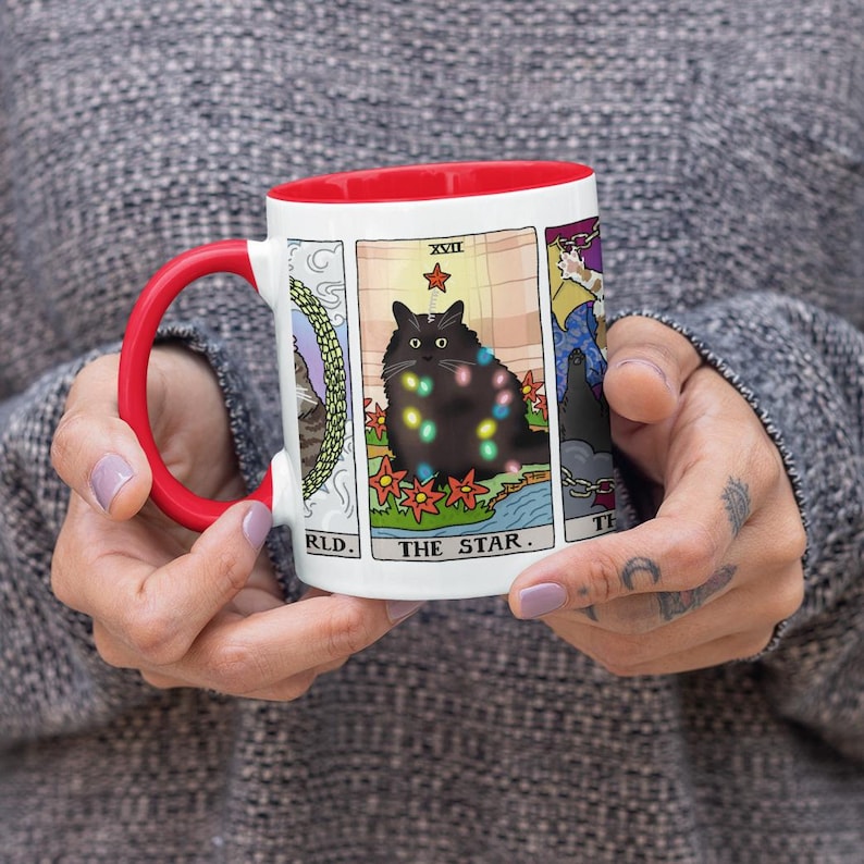 Puede incluir: Taza de cer&aacute;mica blanca con borde rojo, que presenta un gato negro con luces de colores, rodeado de un dise&ntilde;o floral. El gato est&aacute; representado en un estilo de carta de tarot con el texto "The Star" y el n&uacute;mero romano "XVIII" sobre el gato.