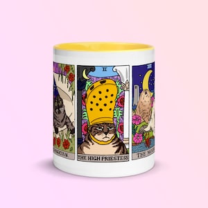 May include: White ceramic mug with a yellow rim. The mug features a design of four tarot cards with cats. The cards are "The Magician", "The High Priestess", "The Chariot", and "The Moon".