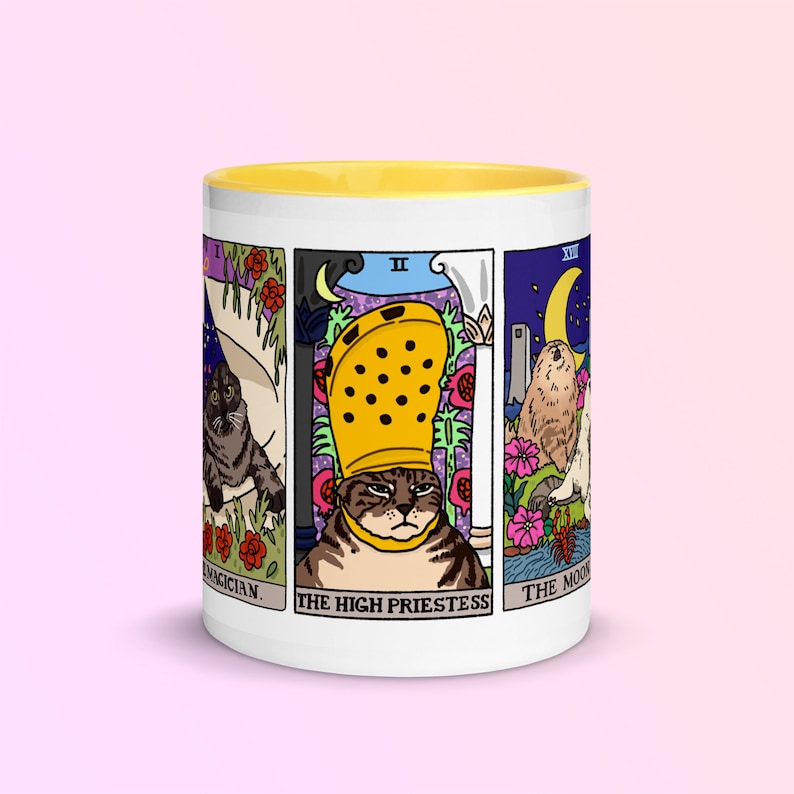 May include: White ceramic mug with a yellow rim. The mug features a design of four tarot cards with cats. The cards are "The Magician", "The High Priestess", "The Chariot", and "The Moon".