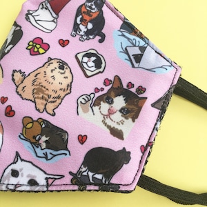 May include: Pink face mask with a repeating pattern of cats and hearts. The cats are in various poses, including sleeping, sitting, and giving a thumbs up. The mask has black elastic straps.