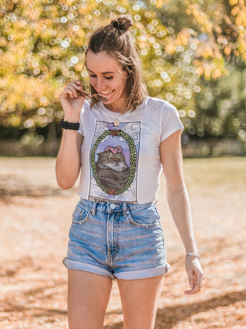 Pu&ograve; includere: Una donna indossa una maglietta bianca con un'illustrazione di un gatto e la scritta "The Cat" stampata su di essa. Indossa anche pantaloncini di jeans e un braccialetto d'argento.