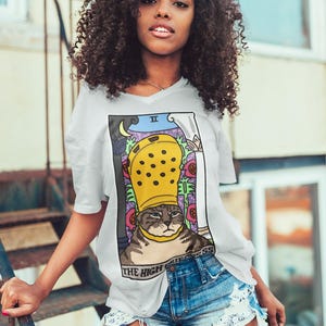 Könnte beinhalten: Weißes V-Ausschnitt-T-Shirt mit einem Tarotkarten-Design mit einer Katze, die einen gelben Clog-Hut trägt. Die Grafik enthält die Worte "THE HIGH PRIESTESS". Das Model trägt Jeansshorts.