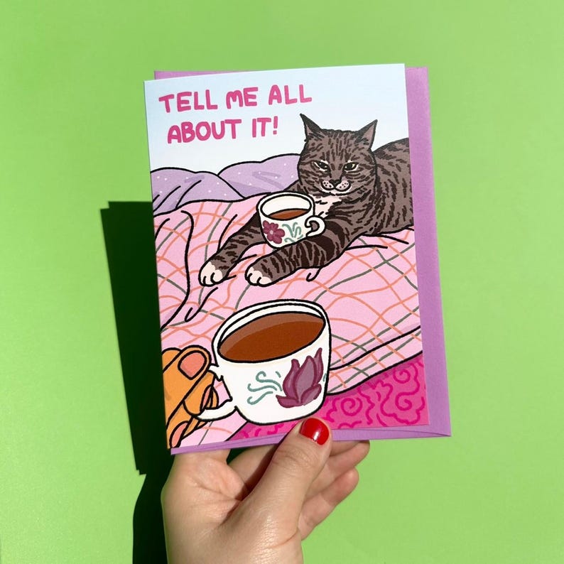 Puede incluir: Una tarjeta de felicitaci&oacute;n con una ilustraci&oacute;n de un gato. El gato est&aacute; acostado sobre una manta rosa y sosteniendo una taza de t&eacute;. La tarjeta dice "TELL ME ALL ABOUT IT!" en texto rosa. Una mano sostiene la tarjeta con una taza de caf&eacute;.