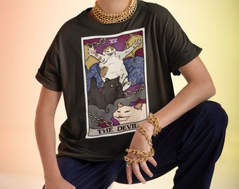Może przedstawiać: Osoba w ciemnozielonym t-shircie z nadrukiem karty tarota z motywem kotów. Karta przedstawia trzy koty i napis "THE DEVIL" w fioletowym polu. Osoba nosi złoty naszyjnik z łańcuszkiem i kilka złotych bransoletek z łańcuszkiem.