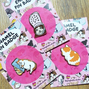 May include: A set of enamel pins featuring various cat designs. The pins are on a pink background with white text that reads "Enamel Pin Badge". The pins include a cat with a crown, a cat with a fish, a cat with a heart, and a cat with a fishbone.