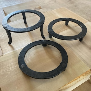 May include: Three black metal trivet stands of different sizes. The trivets are round with a raised edge and three legs.