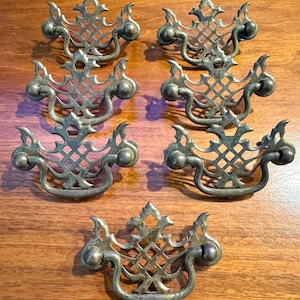 May include: Six antique brass drawer pulls with an ornate, openwork design. Each pull features a decorative center and curved handles, perfect for vintage furniture restoration. The pulls are arranged on a wooden surface, showcasing their intricate details.