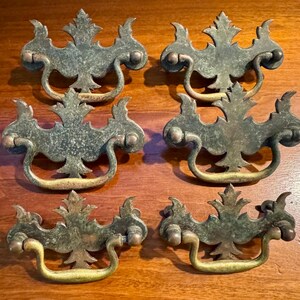 May include: Five antique brass drawer pulls with a leaf design. The pulls have a dark green patina with gold-colored handles and are arranged on a wooden surface. These decorative pulls are ideal for vintage furniture restoration projects.