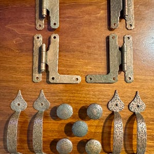 May include: A collection of antique bronze hardware, including hinges, drawer pulls, and knobs. The hinges are L-shaped with a textured finish. The drawer pulls have a decorative, pointed design. The knobs are round and textured.