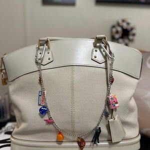 May include: A white leather tote bag with a silver chain strap and multiple charms attached. The charms include a blue dice, a pink dinosaur, an orange ball, a pizza slice, a black figure, a blue eye, a pink box with text, and a silver tag.