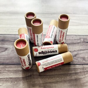 May include: A collection of Sweetlips lip balms in cardboard tubes, with labels featuring the flavors raspberry and chocolate pop. The lip balms are arranged on a wooden surface, with the product's name and flavor clearly visible.