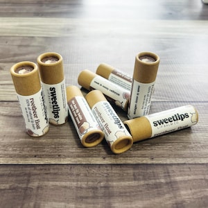 Puede incluir: Varios bálsamos labiales rootbeer float de Sweetlips Lip Balm Co. están colocados sobre una superficie de madera. Los bálsamos labiales están en tubos cilíndricos de cartón con una tapa marrón y una etiqueta blanca. La etiqueta incluye el texto "rootbeer float".