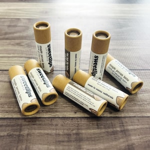 Könnte beinhalten: Auf einer Holzoberfläche liegen mehrere Sweetlips Lippenbalsam-Tuben. Die Tuben haben eine hellbraune Kartonbasis und ein weißes Etikett. Auf den Etiketten steht "maple pecan latte" und "100% all natural lip balm".