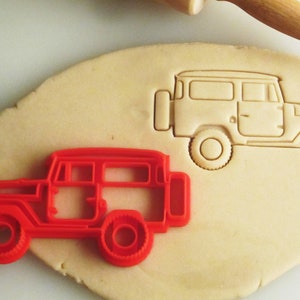 May include: Red plastic cookie cutter shaped like a vintage Land Cruiser. The cutter is on top of a sheet of unbaked cookie dough with an outline of the Land Cruiser pressed into the dough.