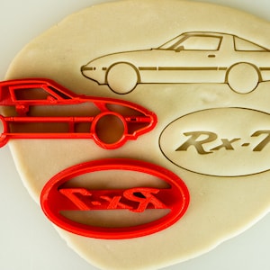 May include: Red plastic cookie cutters shaped like a car and an oval with the text "Rx-7" inside. The cutters are on a beige surface.