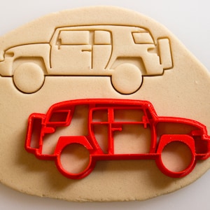 May include: A red plastic cookie cutter in the shape of a car. The cutter is on top of a piece of beige cookie dough.