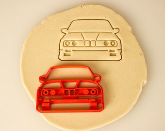 May include: A red plastic cookie cutter in the shape of a car. The cookie cutter is on top of a piece of dough, with a similar cookie cutter in the shape of a car imprinted in the dough.