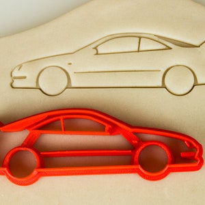 May include: Red plastic cookie cutter shaped like a car. The cutter is on a beige surface with a similar outline of a car.