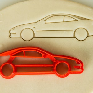 May include: A red plastic cookie cutter shaped like a car. The cookie cutter is on a white surface with a gray outline of the same car.