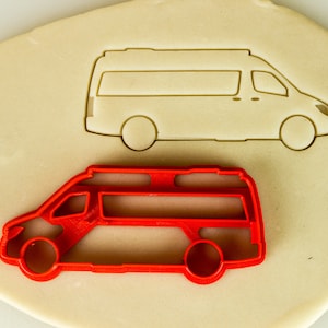 May include: A red plastic cookie cutter shaped like a van with a roof rack. The cutter is on a white surface.