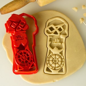 May include: Red plastic skull cookie cutter on top of a piece of unbaked cookie dough. The cookie cutter is shaped like a skull with a crossbones design and a wheel at the bottom.