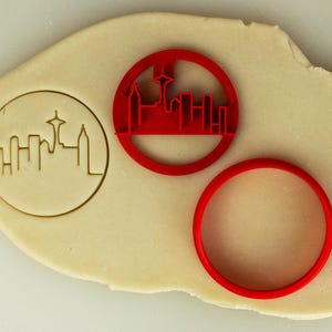 May include: A red cookie cutter depicting the Seattle skyline, including the Space Needle, is next to a matching red circle cutter. The cutters are on a sheet of dough, with an imprint of the skyline visible.