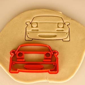 May include: A red plastic cookie cutter shaped like the front of a car. The cookie cutter is on top of a piece of cookie dough.