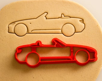 May include: A red plastic cookie cutter shaped like a convertible car. The cutter is on top of a sheet of beige cookie dough.