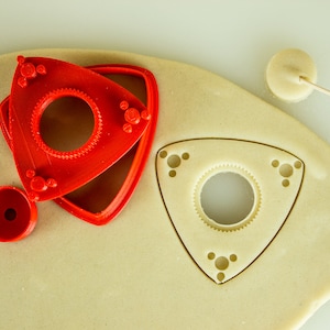 May include: A red plastic cookie cutter in the shape of a triangle with a circle in the center. The cutter is on top of a piece of dough that has been cut out with the same shape. A small, round piece of dough is on the right side of the image with a toothpick sticking out of it.