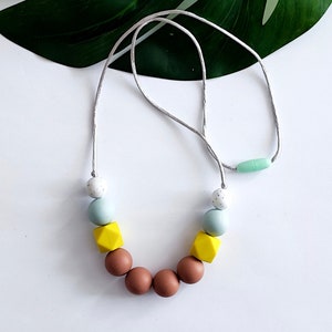 May include: A light gray necklace with a variety of silicone beads in shades of brown, yellow, blue, and white. The beads are round, square, and hexagonal.