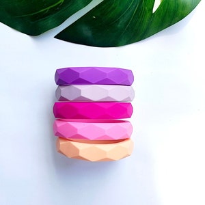 Könnte beinhalten: Ein Stapel aus fünf Silikonringen in einem Farbverlauf von Lila bis Pfirsich. Die Ringe sind mit einem geometrischen Muster strukturiert.