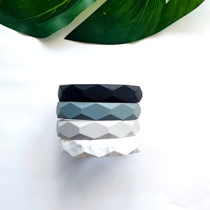 May include: Four stacked rings in shades of black, gray, and white. Each ring has a geometric, faceted design. The top ring is black, followed by gray, light gray, and a marbled white ring. A green leaf is in the background.