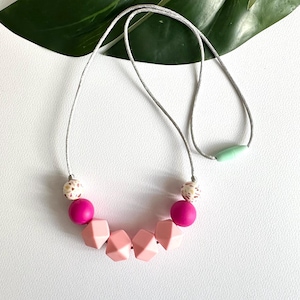 May include: A necklace with a gray cord featuring a variety of beads. The necklace includes pink, light pink, and patterned beads. A light green oval bead is at the back of the necklace. The necklace is on a white surface.